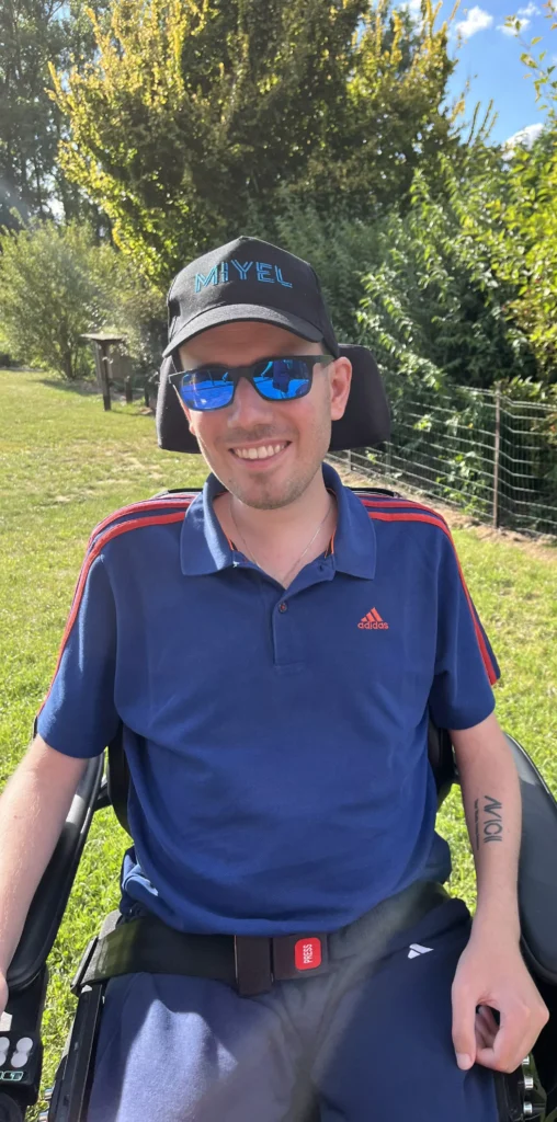 Portrait vom Musiker Michael Großmann. Er sitzt im Rollstuhl, trägt eine blaue Sonnenbrille und lacht freundlich in die Kamera. Er trägt ein Basecap mit seinem Logo. Hintergrund: Draußen im Grünen.
