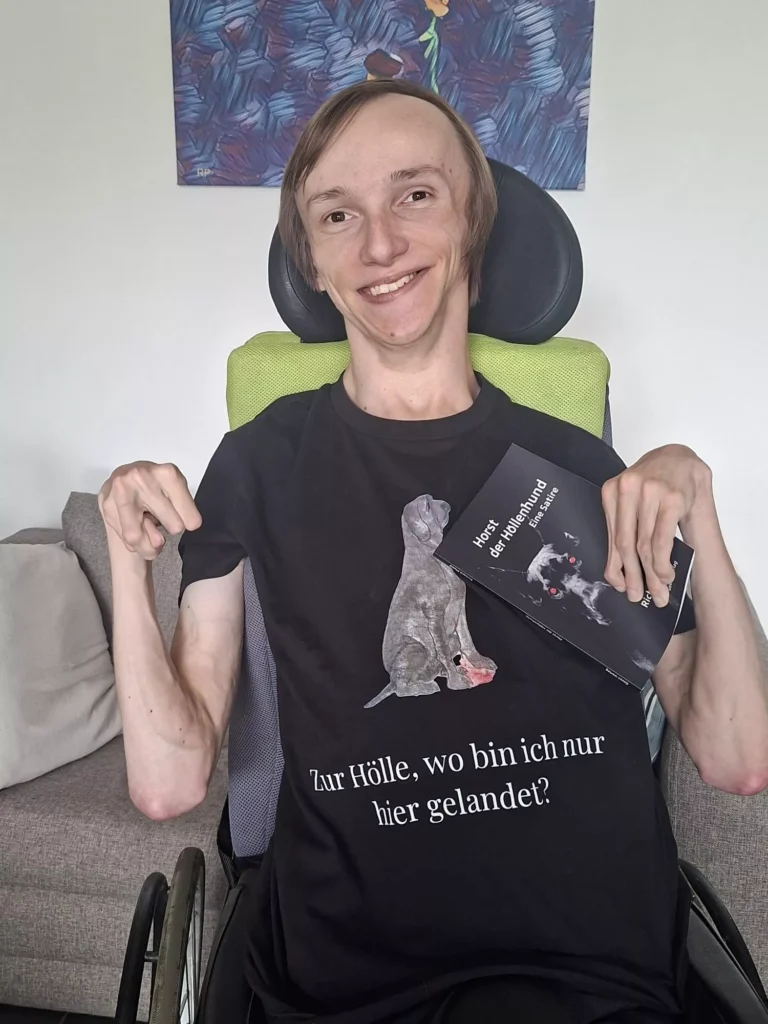 Portrait vom Autor Richard Pfund. Er sitzt im Rollstuhl und lächelt sehr freundlich in die Kamera. Er trägt ein T-Shirt mit einem gezeichneten Hund, Text: Zur Hölle, wo bin ich hier nur gelandet? " In Anlehnung an sein Buch: "Horst der Höllenhund", welches er hier stolz in die Kamera hält.